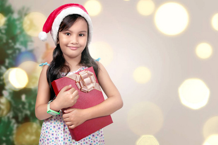 可爱的亚洲小女孩与圣诞老人帽子举行圣诞节礼物盒模糊灯背景