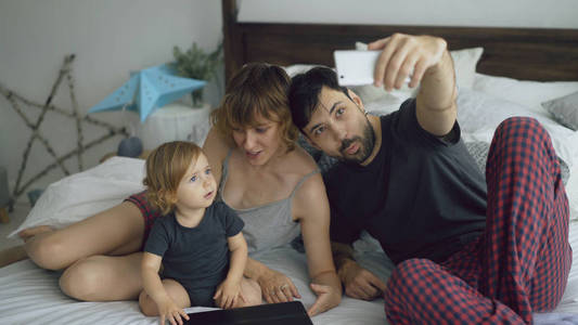 年轻的家庭与可爱的小女孩采取自拍肖像智能手机相机坐在床上在家