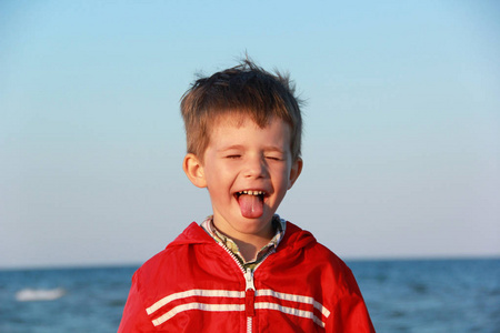 一个开朗快乐的男孩，穿红色夹克做鬼脸和伸出舌头的画像。海边的夕阳