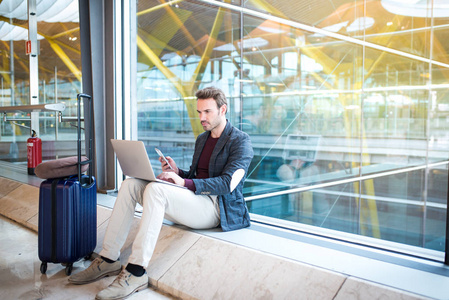 坐在机场使用笔记本电脑和移动电话在窗户旁的男人