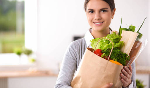 年轻女子捧着与蔬菜站在厨房里的食品杂货店购物袋