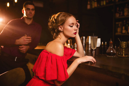 有魅力的年轻女子穿着红裙子坐在柜台上, 背景英俊的男子。在夜总会约会, 有吸引力的情侣在酒吧