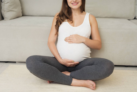 一个孕妇坐在家里的浅色地板上。她坐在地板上, 双腿交叉着, 双手放在肚子上。