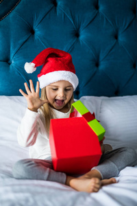 惊喜兴奋的小女孩在圣诞老人的帽子打开目前, 而坐在床上的交叉腿圣诞节早晨