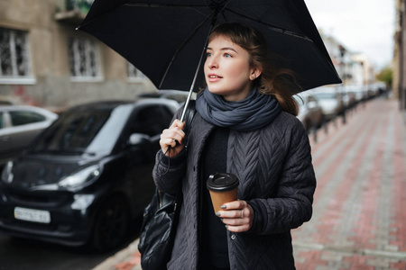 年轻漂亮的女士在街上站着黑色的雨伞和咖啡在手, 朦胧地看着一旁的肖像