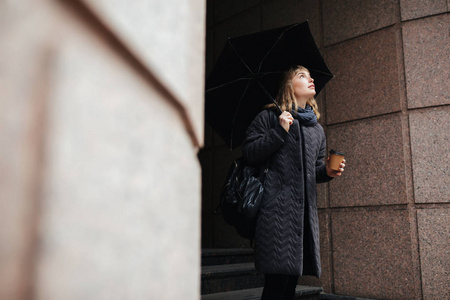 美丽的女士站在街上, 黑色的雨伞和咖啡在手中的肖像, 而朦胧的抬头