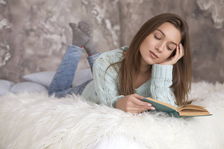 年轻美丽的白种女人坐在床上看书。她穿着休闲服 蓝色毛衣和牛仔裤。长的褐色的头发, 一个小的微笑, 看周到