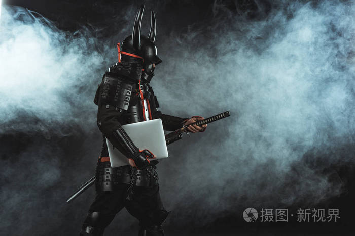 武士的侧面看传统盔甲与膝上型电脑拿出剑黑背景与烟雾