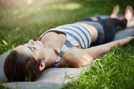 年轻女性冥想和放松在瑜伽垫上在城市公园的草地上在阳光明媚的夏日的特写