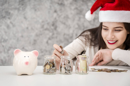 年轻妇女与红色圣诞节帽子 looing 在存钱罐和硬币, 保存概念为圣诞节假日购物