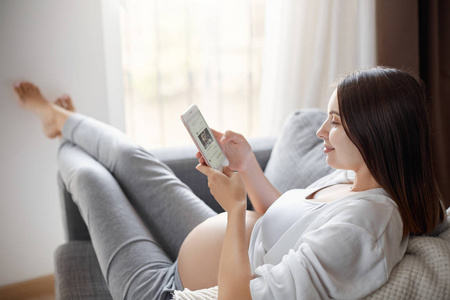 使用智能手机，她在和朋友聊天社交媒体在她舒适的沙发上呆在家里时怀孕少女。积极思考是必须的