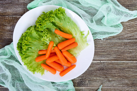 小胡萝卜一个小胡萝卜, 生菜叶子上的白色陶瓷板上的木质背景。适当的营养。健康食品。烹调膳食菜肴的配料