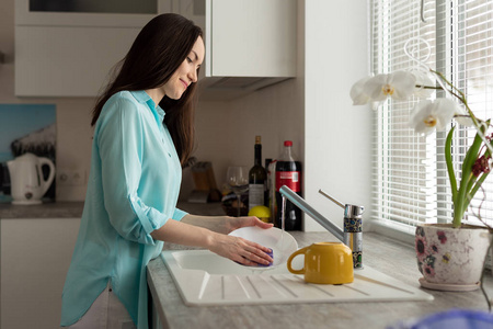 一个穿着绿松石衬衫的女人在厨房的龙头下冲洗盘子。