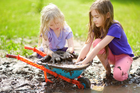 在阳光明媚的夏日, 两个滑稽的小女孩在一个大湿泥水坑里玩耍。孩子们在泥泞的泥土中挖洞时变得肮脏。杂乱的游戏户外