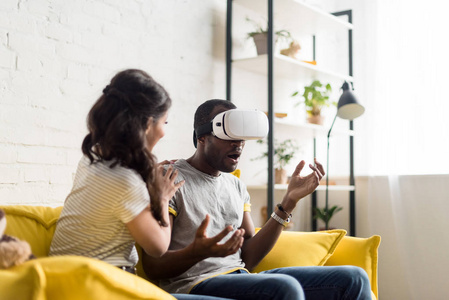 年轻的非洲裔美国妇女坐在附近震惊的男朋友, 而他穿着虚拟现实耳机在家沙发上