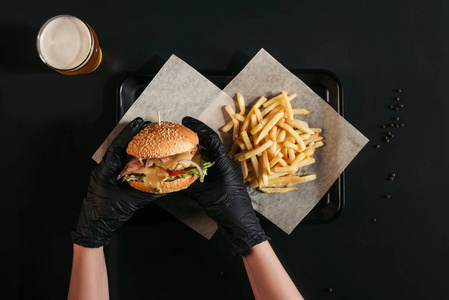 被裁剪的人在手套持有美味的汉堡在托盘上与法式薯条和玻璃啤酒黑色