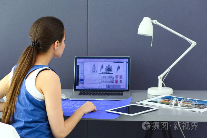年轻的女人坐在办公桌前与文书, 计划和笔记本电脑