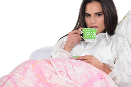 美丽的黑发妇女躺在床上, 拿着一杯咖啡或茶。在白色背景上被隔离