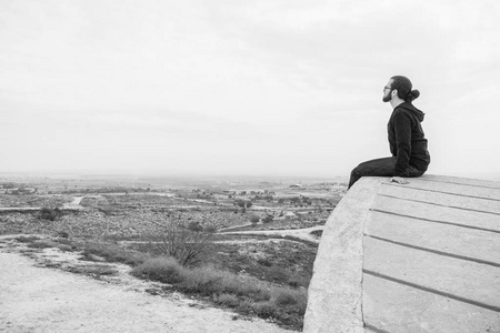 侧面观察不明的年轻人穿着便装和眼镜坐在高高的岩石上, 看着美丽的沙漠。