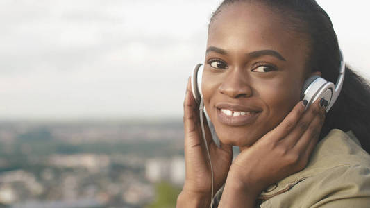 美丽迷人的非洲女孩与自然化妆和可爱的笑容是听音乐在她的耳机在城市的模糊背景