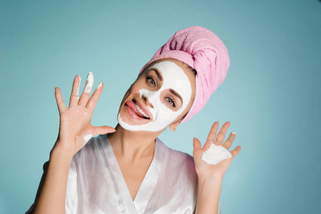 一个滑稽的年轻女孩做面孔, 一条毛巾在她的头, 一个白色保湿面具在她的面孔