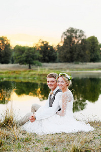 幸福夫妇在婚礼礼服在湖背景日落, 新娘和新郎在白色的礼服