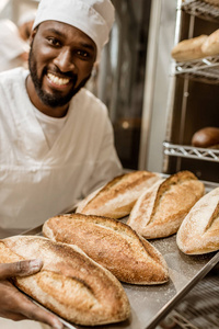 微笑英俊非洲裔美国面包师与盘子新鲜面包托盘在烘烤生产