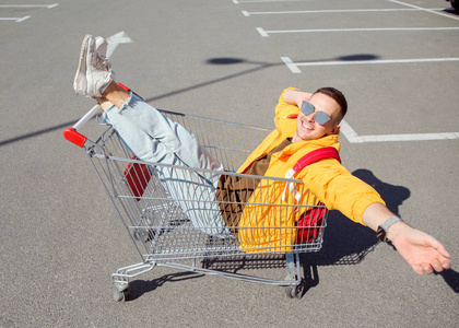时尚的家伙在太阳镜和黄色夹克坐在购物车从食品在超市停车场