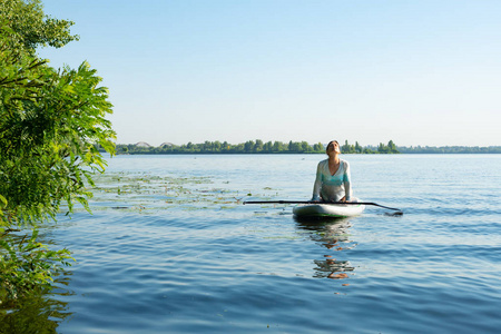 女性在一条大河上的阳光明媚的早晨练习瑜伽。站起来桨登机可怕的活动自然娱乐