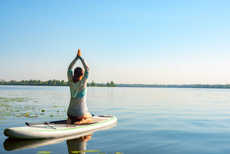 女性在一条大河上的阳光明媚的早晨练习瑜伽。站起来桨登机真棒活跃的娱乐性质。后退视图