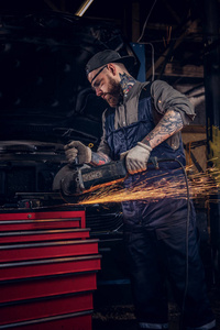 制服和安全眼镜的野蛮的汽车技工在修理车库站在一辆破车上时, 用角磨床工作