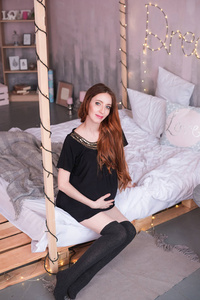 未来的母亲坐在床上。长着红色长发的孕妇, 穿着黑色连衣裙