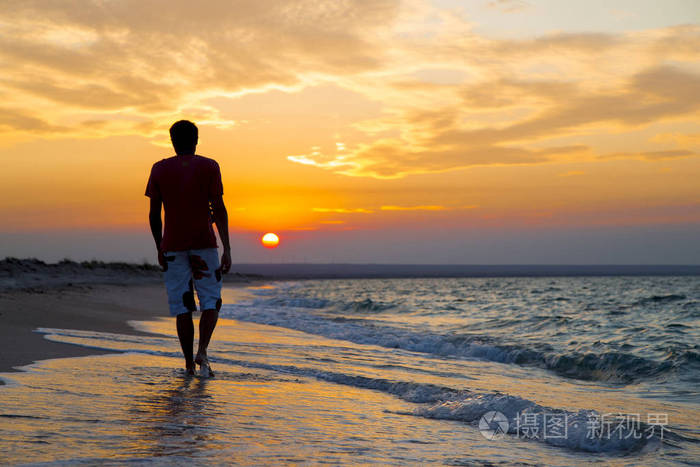 一个年轻男子在海滩上散步的剪影, 在一个美丽的日落的背景下
