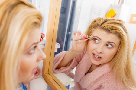 在浴室拔眉毛脱毛用镊子，看着镜子中的女人。钳卸下她面部的头发的女孩。宽视角