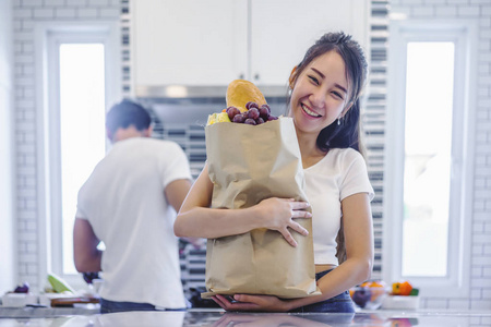 美丽的亚洲年轻夫妇爱笑是期待在厨房做饭在家里, 妇女与购物袋和英俊的男人做饭