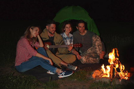 年轻人在晚上篝火旁为朋友弹吉他。野营季节