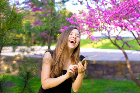 欢乐女孩使用新的智能手机在公园与盛开的树木背景