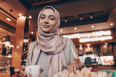 一个可爱的穆斯林女孩戴着头巾坐在一个东方舒适的咖啡馆, 用餐, 看着相机
