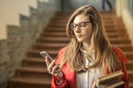 戴眼镜和书本的年轻学生在大学的走廊上检查她的智能手机。