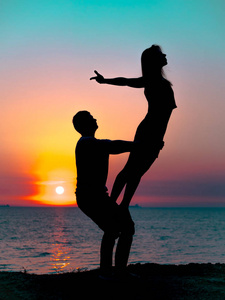 在日落的海边海滩上, 年轻女子和男子做双瑜珈的剪影。冥想。夫妇练习瑜珈。在自然背景下的柔韧性锻炼