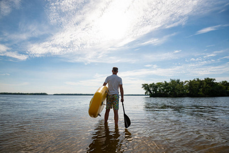 男人沿着海滩散步, 手里拿着一把木板, 双手拿着桨在蓝天背景下行走。站起来桨登机令人敬畏的活动娱乐。后视图, 广角