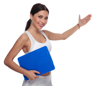 适合的女人欢迎你加入她的健身房与手势, 而持有一个蓝色的剪贴板。她站在白色的背景, 身穿白色的顶部, 肖像图片