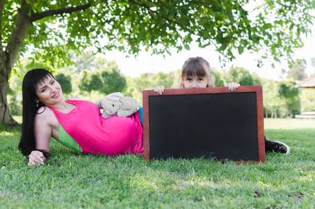 美丽的孕妇躺在公园与她的女儿谁持有空黑板在前面