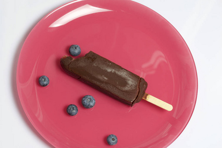 巧克力上的无骨冰淇淋冰在棍子上。躺在一块珊瑚色的盘子里。蓝莓就在他们旁边。白色背景