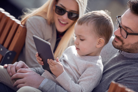 幸福的家庭和数字技术。年轻的父母和小孩坐在城市公园里使用平板电脑