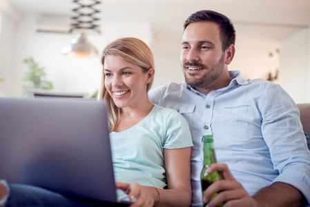 年轻幸福夫妇坐在沙发上的照片在家里。看着开心, 用笔记本电脑