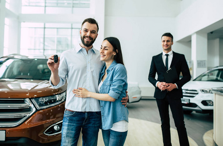 快乐的年轻家庭站在汽车钥匙在经销商, 男性经理和汽车的背景