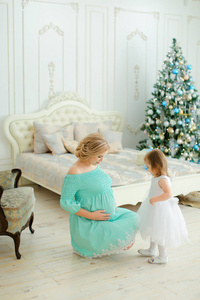 孕妇身穿蓝色连衣裙拥抱腹部, 坐在圣诞树附近的小女儿在卧室