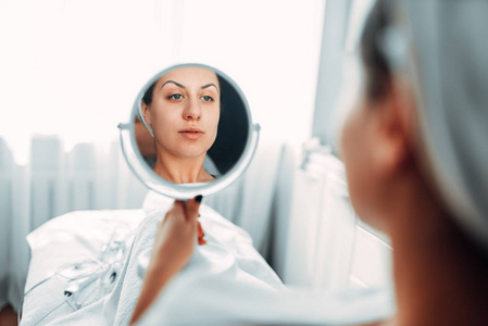 女病人透过镜子, 美容诊所看她的脸。恢复过程的结果。水疗沙龙面部护肤, 美容医学