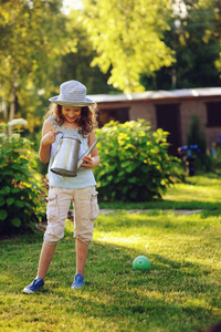幸福的女孩的垂直的画像在园丁帽子玩浇水罐头在阳光明媚的夏日花园